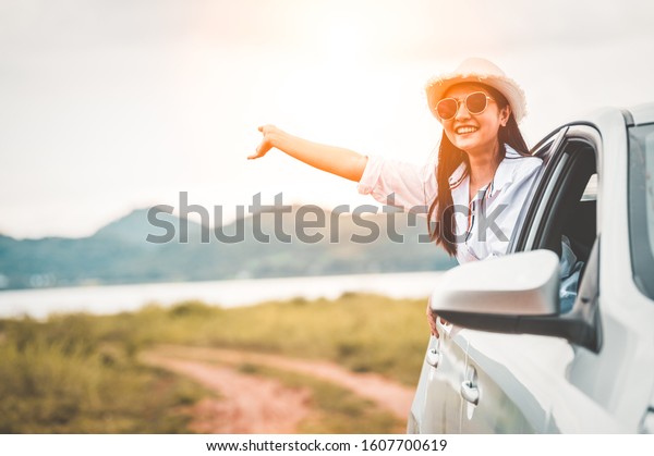 Happy woman waving hand outside open window car\
with meadow and mountain lake background. People lifestyle relaxing\
as traveler on road trip in holiday vacation. Transportation and\
travel concept