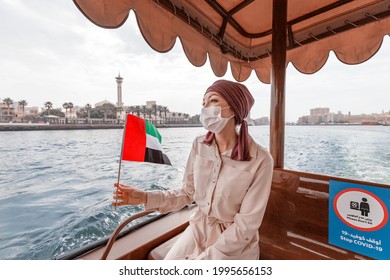 Happy woman wearing a medical mask with the flag of the UAE is traveling on the traditional boat ferry Abra Dhow on the Dubai Creek. Coronavirus and covid-19 are not a hindrance to travel - Shutterstock ID 1995656153