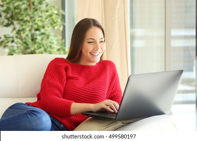 Happy Woman Wearing A Red Sweater Using A Laptop On Line Sitting On A Sofa In The Living Room At Home In Winter