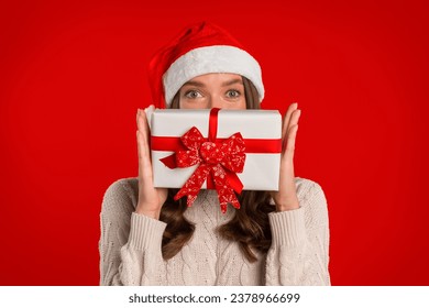 Happy woman wearing santa hat holds Xmas gift box before face on red studio backdrop, looking at camera hiding behind Chistmas present. Festive sales and surprises