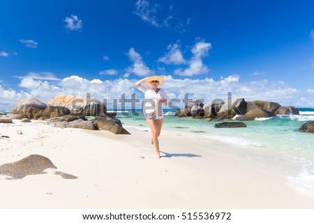 Happy woman wearing white loose tunic over bikini and beach hat, enjoying amazing white sandy beach on Mahe Island, Seychelles. Summer vacations on picture perfect tropical beach concept.