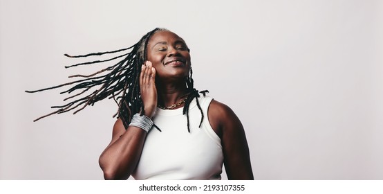 Happy woman whipping her dreadlocks while listening to vibrant music on earbuds. Cheerful mature woman enjoying her favourite playlist while standing against a studio background.