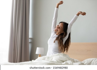 Una mujer feliz con ropa de noche blanca sentada en la cama despertada de suficiente y el sueño saludable se siente bien, estirando los músculos de los brazos después del sueño y la larga inmovilidad despierta empezar un nuevo día con el concepto de sonrisa