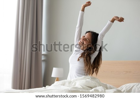 Happy woman in white nightwear sitting in bed awakened from enough and healthy sleep feels good, stretching her arms muscles after sleep and long immobility wakes up start new day with smile concept