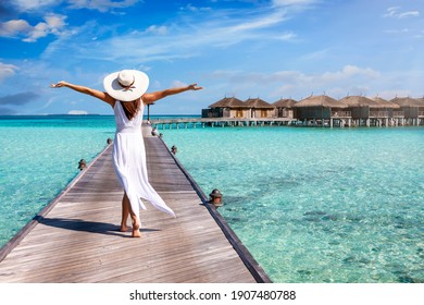 A happy woman in white summer dress and with raised arms walks over a wooden pier in the Maldives islands and enjoys her tropical holidays