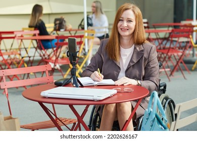 Happy Woman Who Uses A Wheelchair Using Smartphone For Work, Remote Learning, Making Notes In Notebook.