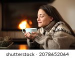 Happy woman in winter drinking coffee beside a fireplace heating at home