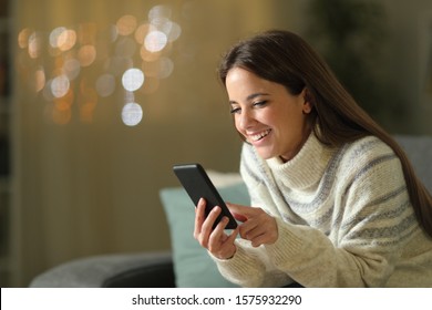 Happy Woman In Winter Using Mobile Phone In The Night Sitting On A Couch In The Living Room At Home