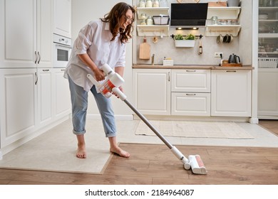 Happy woman with a wireless portable vacuum cleaner in the kitchen. A smiling woman cleans the floor in an apartment with a vacuum cleaner with a battery