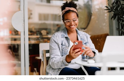 Happy woman works remotely in a café, using her smartphone for communication and social media. Her authentic smile reflects her happiness as a freelancer in the digital age. - Powered by Shutterstock