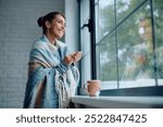 Happy woman wrapped in a blanket enjoying in view from the window while having a cup of tea. Copy space.