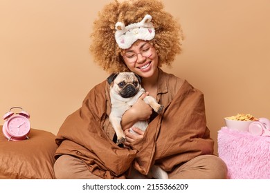 Happy woman wrapped in blanket holds pug dog expresses love to favorite pet has glad expression poses on comfortable bed alarmclock bowl of cornflakes around beige background. Time to having rest