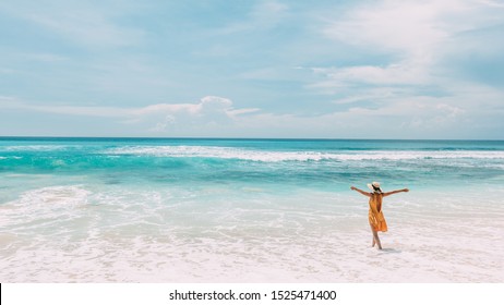 Happy Woman In Yellow Dress Enjoys Her Tropical Beach Vacation While Walking Along Sandy Lagoon With A Paradise View