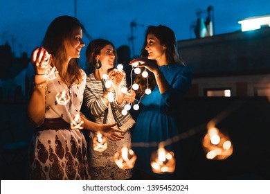 Happy women having fun at rooftop party with lights at night - Three beautiful women wearing summer clothes talking and dancing - Lifestyle and friendship in Barcelona