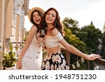 Happy women holding champagne glasses and smiling on balcony. Girl in floral skirt with glass of wine laughts with her friend in wide-brimmed hat