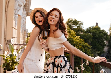 Happy Women Holding Champagne Glasses And Smiling On Balcony. Girl In Floral Skirt With Glass Of Wine Laughts With Her Friend In Wide-brimmed Hat