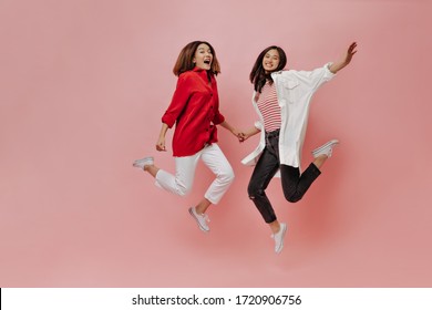 Happy Women Jump On Isolated Background. Tanned Asian Girls In Stylish Outfits Move On Pink Backdrop.