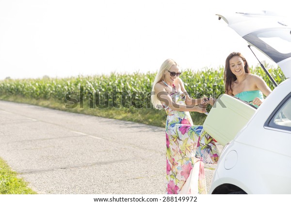 Happy women loading luggage in car trunk against\
clear sky
