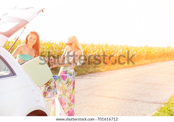 Happy women loading luggage in car trunk against\
clear sky