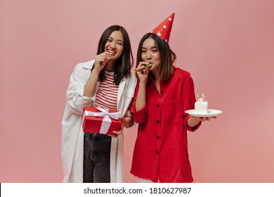 Happy Women In Stylish Outfits Blow Out Party Horns And Laugh. Attractive Asian Brunette Woman Holds Gift Box On Pink Background. Young Girl In Red Blouse Poses With Piece Of Birthday Cake.