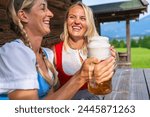 Happy women in traditional dirndls or tracht clinking beer mugs in front of the bavarian alpine mountains at a alpine cabin. Oktoberfest Bavarian german concept image