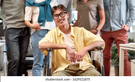 I Am So Happy To Work Here. Portrait Of Young And Positive Asian Man In Wheelchair Looking At Camera And Smiling While His Colleagues Standing In The Background. Disability Concept. Teamwork Concept