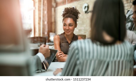 Happy To Work Here. Young And Cheerful Afro American Woman Smiling And Looking Aside While Having A Meeting With Colleagues In The Modern Office