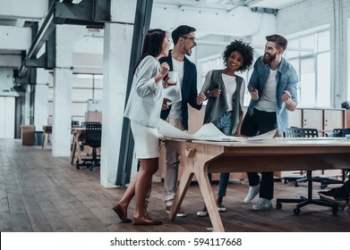 Happy To Work Together. Group Of Young Business People Talking And Smiling While Standing Near The Wooden Desk In The Office