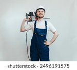 Happy worker holding power drill, wear white shirt and blue overalls, handyman, construction tools, power tools, plain background, drilling, safety gear, industrial equipment, drilling services