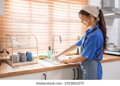 Happy working lifestyle worker concept, portrait housekeeper woman washing dishes in kitchen with natural light from window, Asian cleaning service worker female wearing apron concentrated working - Powered by Shutterstock