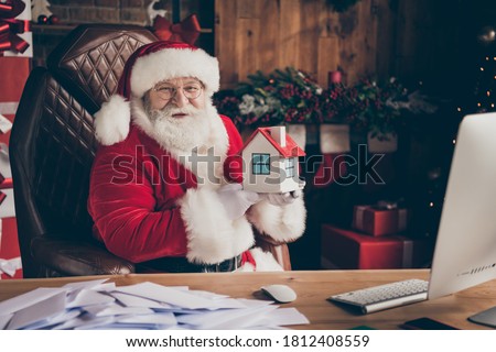 Happy x-mas christmas season shopping sales. Jolly white grey beard hair santa claus sit table hold small insurance building wear cap in house indoors with spirit atmosphere advent tinsels
