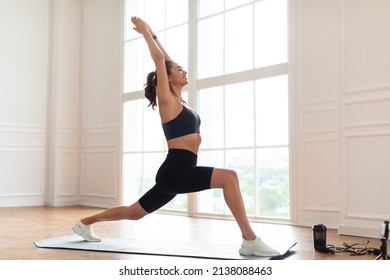 Happy Yogi Woman In Sportswear Exercising Near Window, Standing In Warrior 1 Pose On Fitness Mat Watching Video Using Phone On Tripod, Free Copy Space, Full Body Length. Healthy Sporty Lifestyle