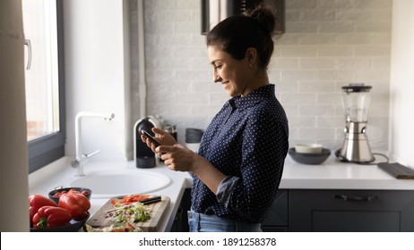 Happy Young 20s Indian Woman Cook Healthy Vegetarian Salad At Home Kitchen Read Recipe On Cellphone. Smiling Ethnic Millennial Female Prepare Diet Food Use Smartphone Texting Or Messaging.