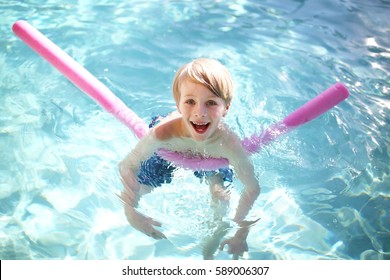 A Happy, Young, 7 Year Old Boy Child Is Smiling As He Takes Swimming Lessons With A Floating Fun Noodle Outside In The Pool On A Summer Day.