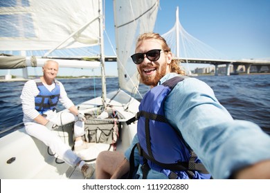 Happy Young Active Man In Sunglasses And Lifejacket Making Selfie During Sailing With Senior Friend