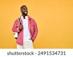 Happy young adult African man hipster standing isolated on yellow background looking away. Smiling generation z Black student guy wearing backpack headphones advertising travel or education offers.