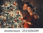 A happy young adult couple in love in an affectionate embrace, celebrating Christmas and decorating the Christmas tree.