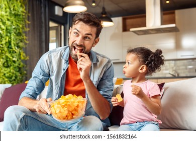 Happy Young Adult Father Sitting On Comfort Couch With His Cute African Child Girl. Family Spending Day Together At Home, Eating Chips And Watching Tv