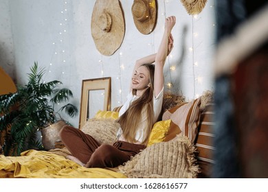 Happy Young Adult Girl In Nightwear Resting In Bedroom, Spending Morning At Home, Sitting On Bed, Raised Hands Up And Smiling Wide