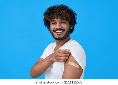 Happy Young Adult Indian Latin Man Showing Bandage Plaster On Arm After Getting Vaccination Isolated On Blue. Vaccine And People Inoculation, Immunization For Covid Prevention Concept. Portrait