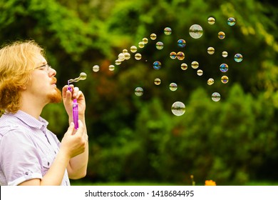 Happy young adult man blowing soap bubbles in nature green spring park.