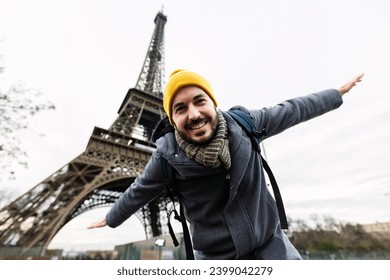 Happy young adult man enjoying travel vacation in Paris. Joyful european tourist male opening his arms pretending to be an airplane in front of Eiffel Tower. Holiday and travel concept.