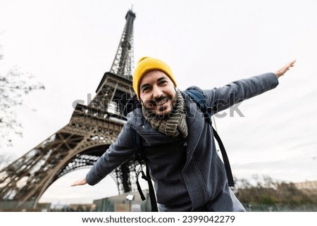 Happy young adult man enjoying travel vacation in Paris. Joyful european tourist male opening his arms pretending to be an airplane in front of Eiffel Tower. Holiday and travel concept.