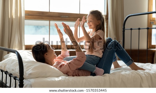 Happy young adult mommy and cute kid daughter play\
patty cake in bedroom, playful family mother with little child girl\
having fun clap hands together laugh enjoy funny sunny morning game\
relax on bed