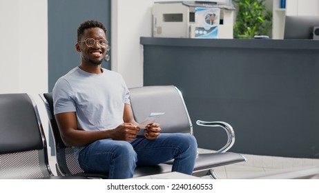 Happy young adult waiting in hospital lobby to meet with general practitioner, sitting in waiting room before consultation. Smiling patient in front desk reception area having appointment. - Powered by Shutterstock