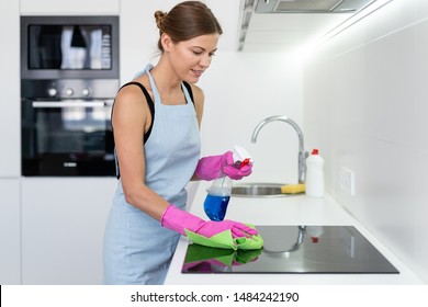 Happy young adult woman in apron and rubber gloves, cleaning induction stove in modern kitchen, using wipe cloth and detergent spray