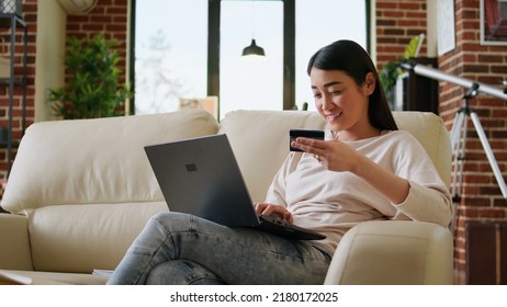 Happy Young Adult Woman Inserting Credit Card Credentials On On Shopping Site For Online Payment. Asian Customer Sitting On Couch At Home While Paying For Products On Digital Store.