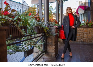 Happy young adult woman smiling with teeth smile, walking outdoors on city street at sunset time. Attractive girl weared in autumn clothes. Red bag, black coat. Blurred details