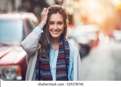 Happy young adult woman smiling with teeth smile outdoors and walking on city street at sunset time weating winter clothes and knitted scarf.