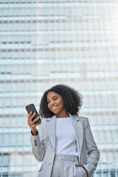 Happy Young African American Business Woman Using Cell Phone Outdoor. Smiling Lady Professional Holding Smartphone Standing At City Street Building Looking At Mobile Working Outside Office, Vertical.